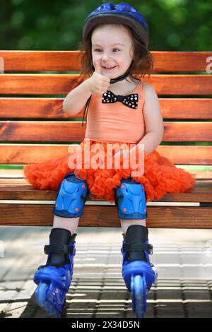 Girl in roller skates sits on bench and thumps up at summer day Stock Photo