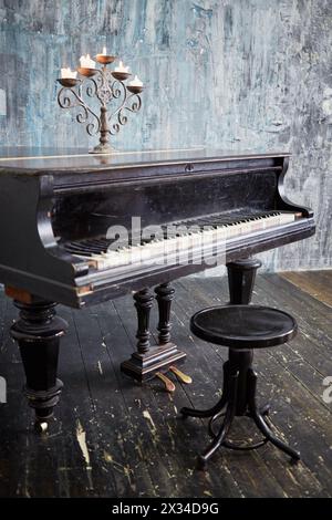 Old black grand piano with burning candles in candlestick at lid stands in room with ragged walls and floor. Stock Photo