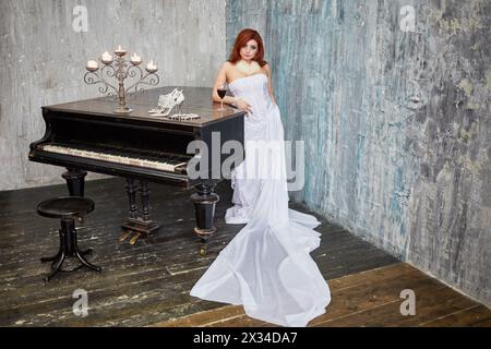 Red-haired woman in white dress stands leaning her elbow on grand piano lid with glass of red wine, burning candles, shoes and necklace in room with r Stock Photo