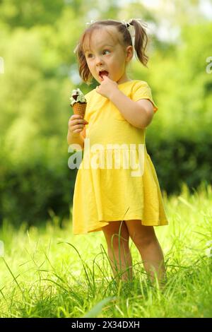 Cute little girl in yellow dress eats sweet ice cream in green park Stock Photo