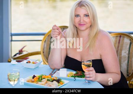 Blonde woman sitting at table in ship restaurant, eating and drinking wine Stock Photo