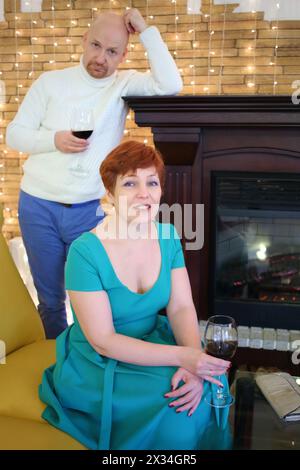 Portrait of a woman in a green dress and a man in a white sweater with glasses of wine near the fireplace Stock Photo