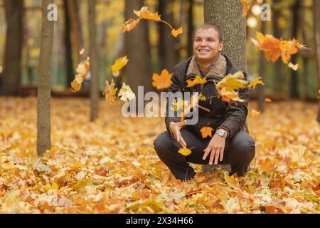 young man in black jacket in autumn park, crouched near tree, throws up yellow autumn leaves, smiling Stock Photo