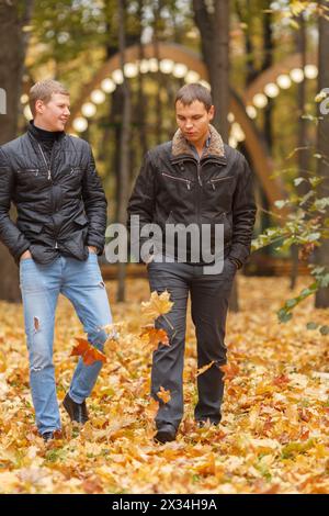 two young man in black jacket in autumn park walking on yellow leaves, one of them smiling Stock Photo