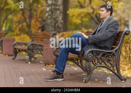 portrait of young man in gray coat and jeans sitting on bench in alley in park, side view Stock Photo