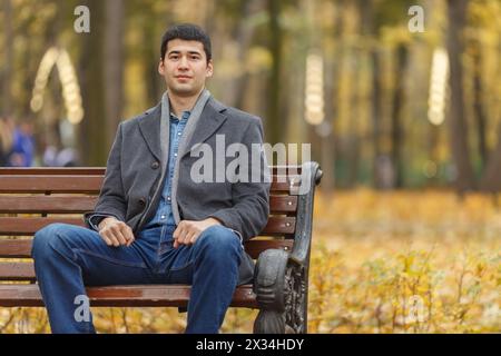 portrait of young man in gray coat and jeans sitting on bench in alley in autumn park Stock Photo