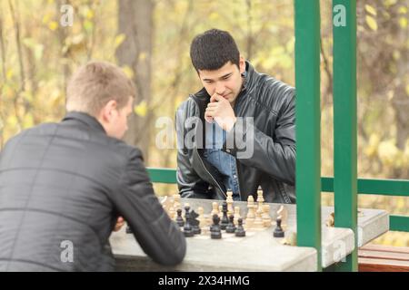 Two young men in black jackets playing chess outdoors in park Stock Photo