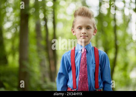 Portrait of boy wearing red trousers with braces, blue shirt, red tie in summer park. Stock Photo