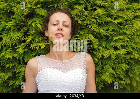 half length portrait of beautiful woman in white dress in summer park on background of green bush Stock Photo