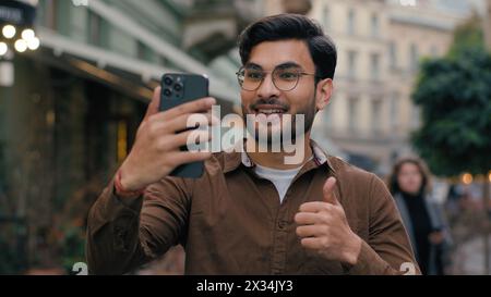 Happy smiling Indian Arabian ethnic man male student gen z guy blogger using modern cellphone recording video travelling blog mobile phone waving hand Stock Photo