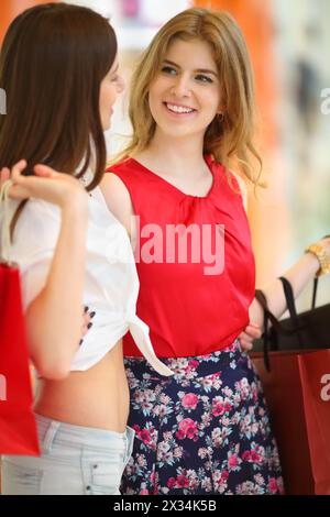 Two pretty girls stand near showcase with bags and look at each other in shopping center. Focus on right woman Stock Photo