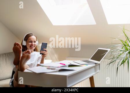 Carefree female student sits at the desk at home and chatting online using her smartphone. Stock Photo