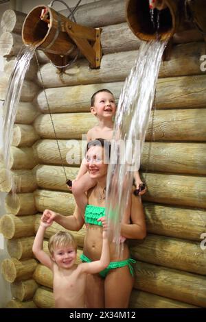 Happy woman with two boys overturns the wooden buckets with water near log wall in a sauna Stock Photo