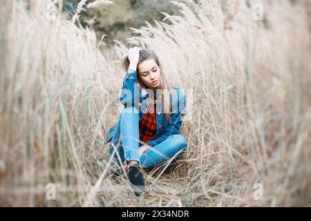 Pretty Girl Sits In A Beautiful Autumn Grass. Blue JeanYoung Beautiful Girl With A Smile In Denim Clothes. Jacket. Stock Photo