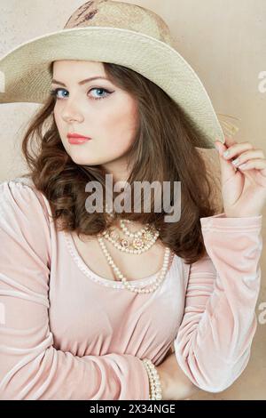 Young woman in pink dress with pearl necklace, beads and wide-brimmed hat on head at wall in studio. Stock Photo