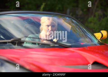 Blonde pretty woman sits in red sport car looks at camera at sunny day Stock Photo