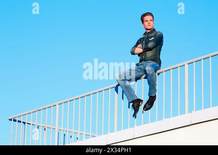 Young man in denim clothes sits with his arms crossed on his chest, on the metal railing of the building roof. Stock Photo