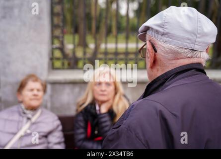 A senior man wearing a hat talking in front of two women sitting in a park. The scene appears to be casual and friendly Stock Photo