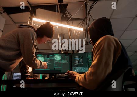 Hackers team hacking online server, stealing data, cybercrime. Cyberattack, it specialist pointing at code on pc screen, criminals programming internet virus at night time Stock Photo