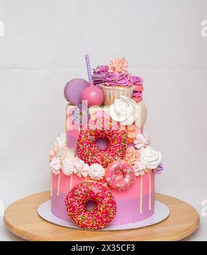 Pink ombre birthday cake with donuts, meringues and chocolate on neutral background. Cupcake on the top Stock Photo