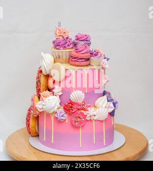 Pink ombre birthday cake with donuts, meringues and chocolate on neutral background. Cupcake on the top Stock Photo