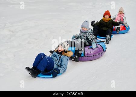 Row of adorable happy children in winter jackets sitting on slides and snow tubes and riding down hill in natural environment Stock Photo