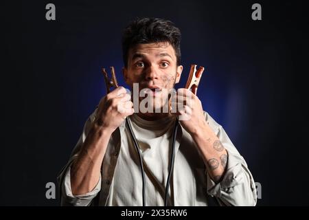 Electrocuted young man with burnt face and jumper cables on dark background Stock Photo