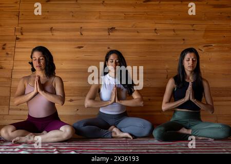 Front view of three young Latin American women meditate together in yoga class. Background of wooden slats. Concept of wellness and yoga. Stock Photo