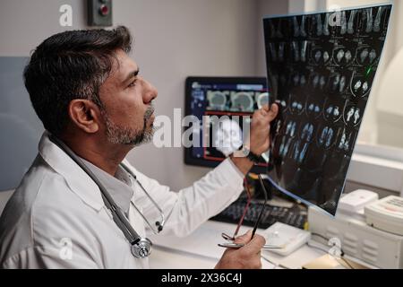 Mature male radiographer sitting at table in office working with head X-ray scan, medium closeup Stock Photo