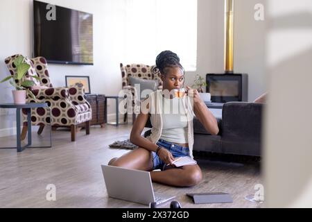 African American woman sits on the floor, sips juice, and looks at a laptop Stock Photo
