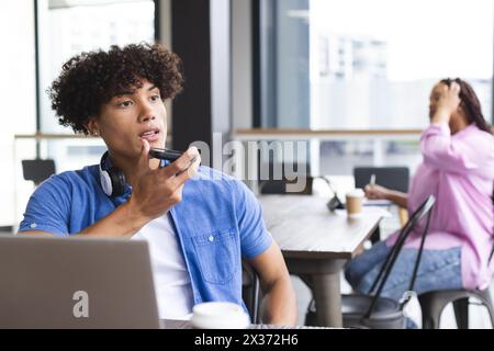 Biracial man with headphones talks on a smartphone, woman nearby in modern office Stock Photo