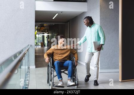 African American man walks beside smiling biracial man in wheelchair in a business office Stock Photo