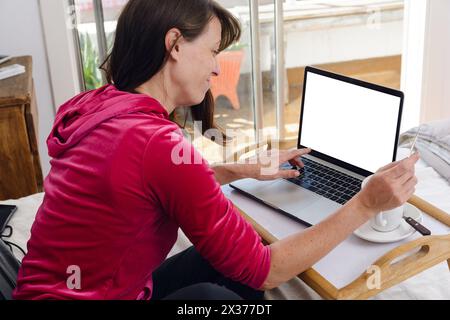 rear view of latin adult woman in pajamas at home sitting on bed in her room, with laptop making online payment with her credit card to watch movies o Stock Photo