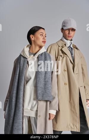 A young stylish couple wearing trench coats and hats in a studio setting against a grey background. Stock Photo