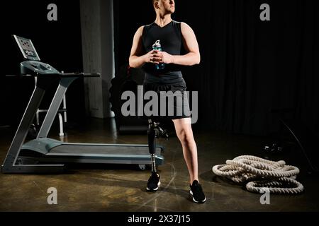 A disabled man with a prosthetic leg stands in front of a treadmill, holding a water bottle in a gym setting. Stock Photo