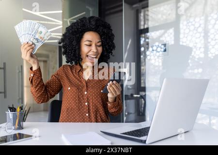 Enthusiastic African American businesswoman holding a fan of cash and excitedly looking at her mobile phone in a modern office setting. Expression of financial success and joy. Stock Photo