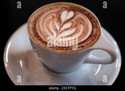 White cup on saucer with trendy latte art coffee. On black background. Perfect cappuccino. Floral pattern. Top view, focus on the foam Stock Photo