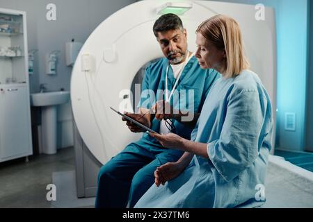 Biracial radiographer sitting on CT scanner bed showing X-ray image to mature Caucasian female patient, copy space Stock Photo