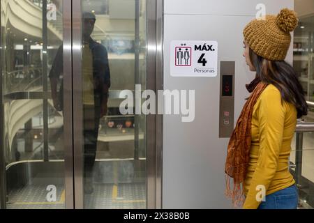 Young Latin American woman in the mall waits for the doors to open to enter the elevator. Dressed in yellow cap and long sleeve flannel. Concept movin Stock Photo