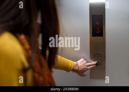 Unrecognizable young Latin American woman (23) in a shopping mall presses the elevator button. She is wearing yellow long-sleeved flannel. Concept of Stock Photo