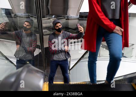 Young man (22) Latin American with face mask goes up the escalator in a shopping mall and takes a self-portrait with his cell phone Concept walk and l Stock Photo