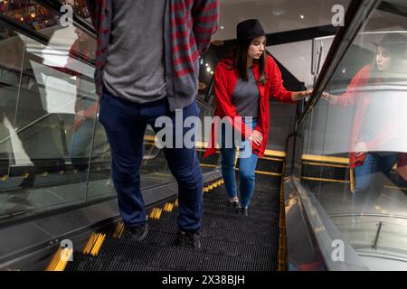 Young Latin American woman (23) walks up the escalator in a shopping mall. She is wearing a black hat and red sweater. Walking and leisure concept. Stock Photo