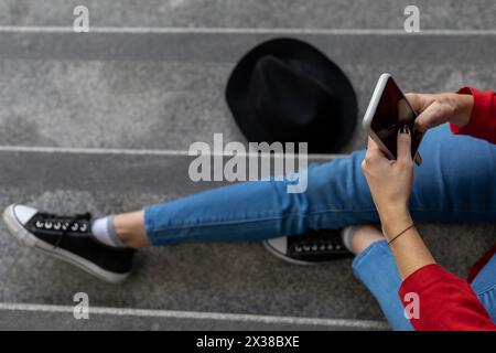 Young (23) unrecognizable Latin American woman (23) sitting on the stairs checking her cell phone. View from above. Technology concept. Stock Photo