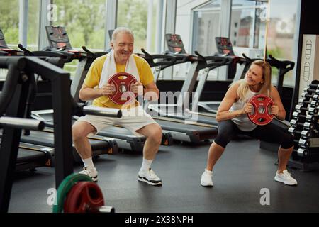 Fit elderly couple, man and woman smiling of joy during strength training, lifting weight plates in modern gym. Stock Photo