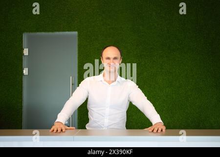 Smiling man in white shirt stands behind desk of reception with green wall Stock Photo