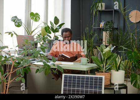 Portrait of adult African American man reading notes in planer while sitting at workplace with lush green plants copy space  Stock Photo