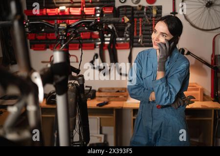 Female mechanic talking on mobile phone. Girl calls after fixing bike in atmospheric garage Stock Photo