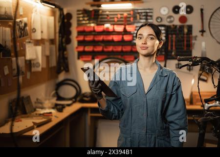 Happy female supervisor mechanic with cheerful smile, inspects repair with tablet at garage, service bike maintenance Stock Photo
