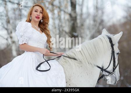 Young woman in white dress rides on white horse in park. Stock Photo