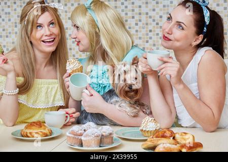 Three young women gossipers and little dog sit at table with baked sweets in kitchen. Stock Photo